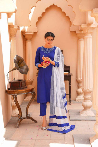 Woman in a blue traditional outfit with a white dupatta in an ornate indoor setting.